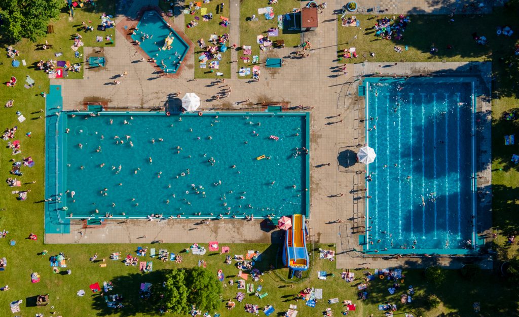 Aerial view of a two large and crowded outdoor pool. People swim in blue water pool top view on a summer day. Pool in green park.